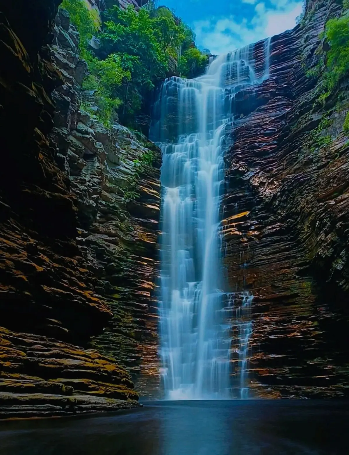 Cachoeira do Buracão: queda d'água alta e fina entre paredões de rocha em camadas, caindo num poço escuro sob o céu azul