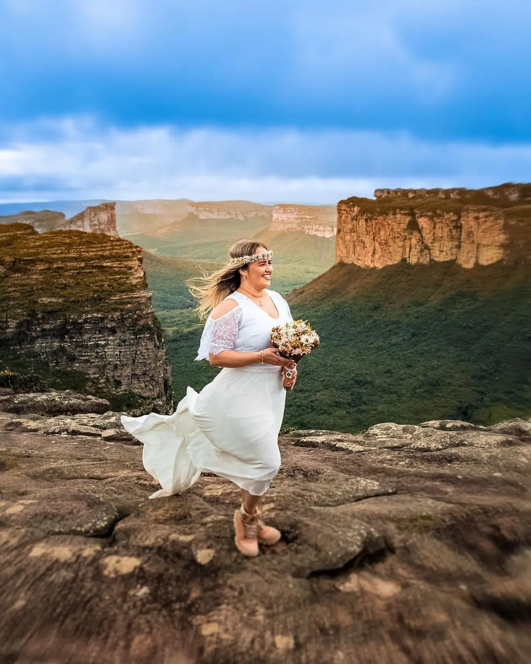 Noiva de vestido branco, coroa de flores e buquê, sorrindo no topo do Morro do Pai Inácio com o vento no cabelo, diante de morros de topo plano e do vale verde