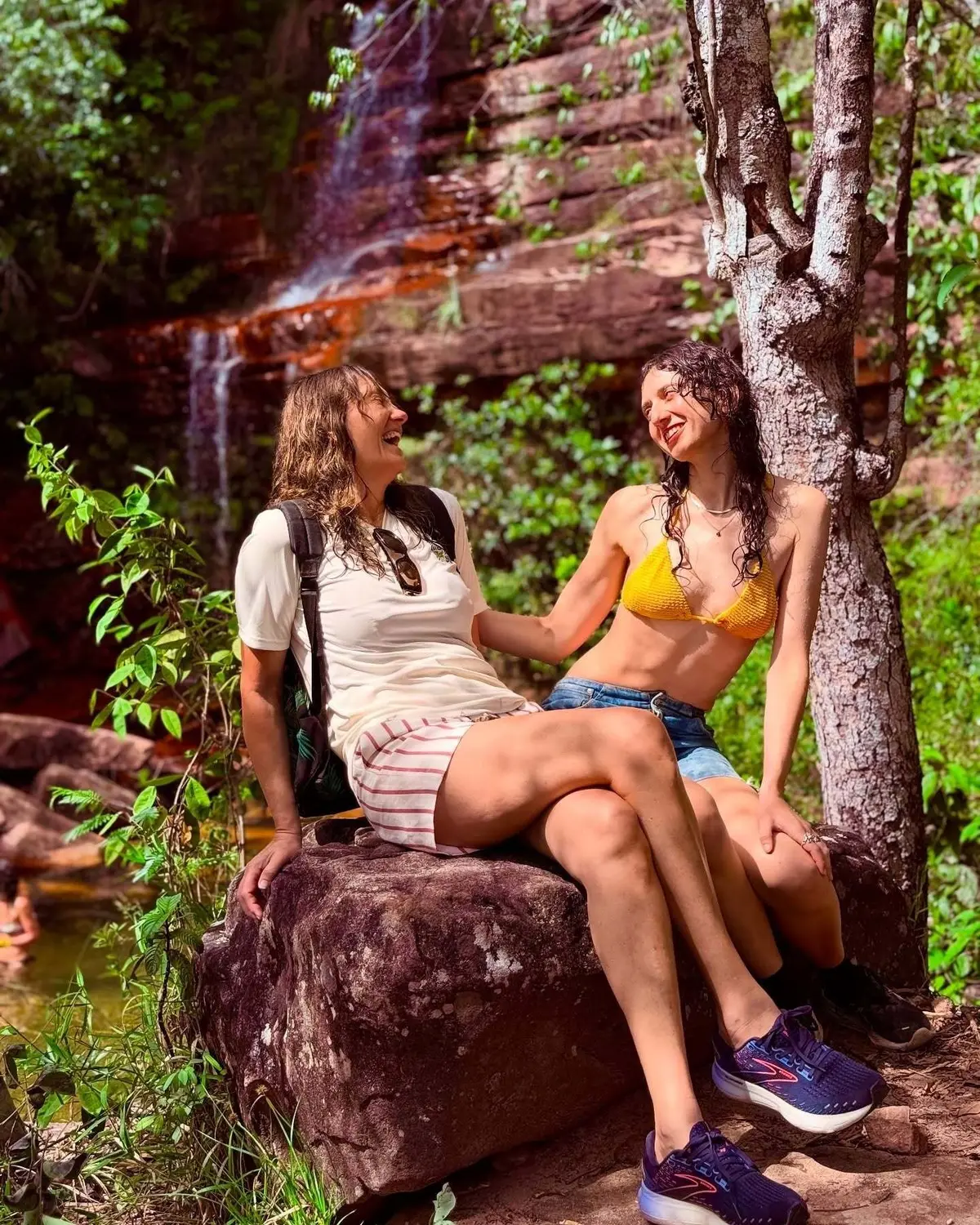 Duas amigas rindo sentadas numa pedra, com quedas finas sobre arenito avermelhado ao fundo