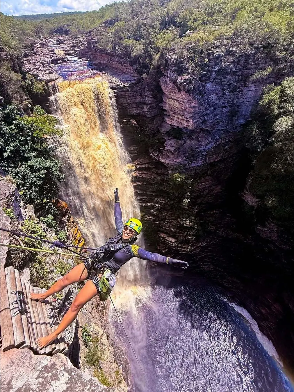 Mulher de capacete fazendo rapel de braços abertos ao lado da cachoeira do Poço do Diabo