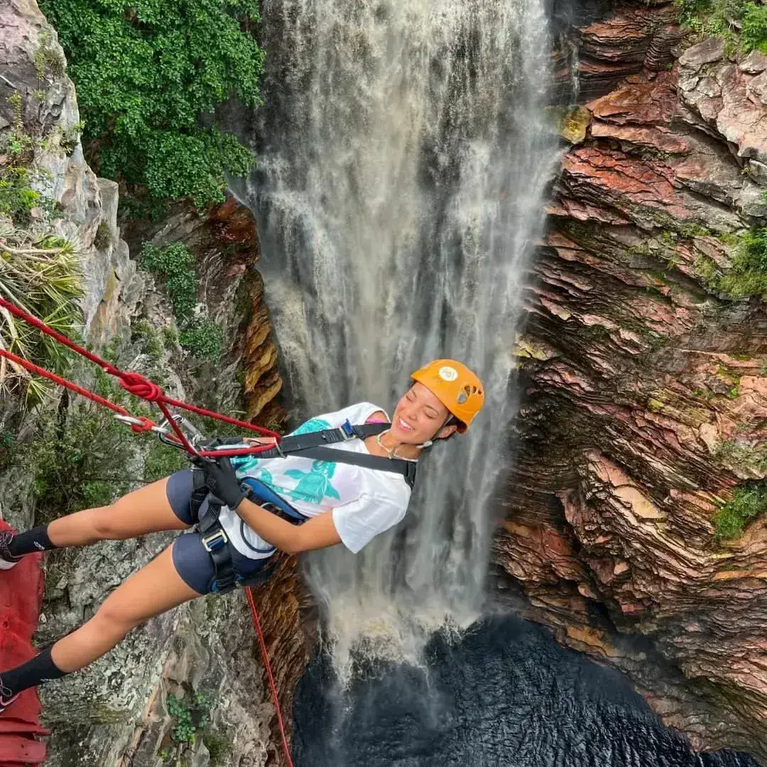 Moça de capacete laranja sorrindo no rapel ao lado da cachoeira do Poço do Diabo