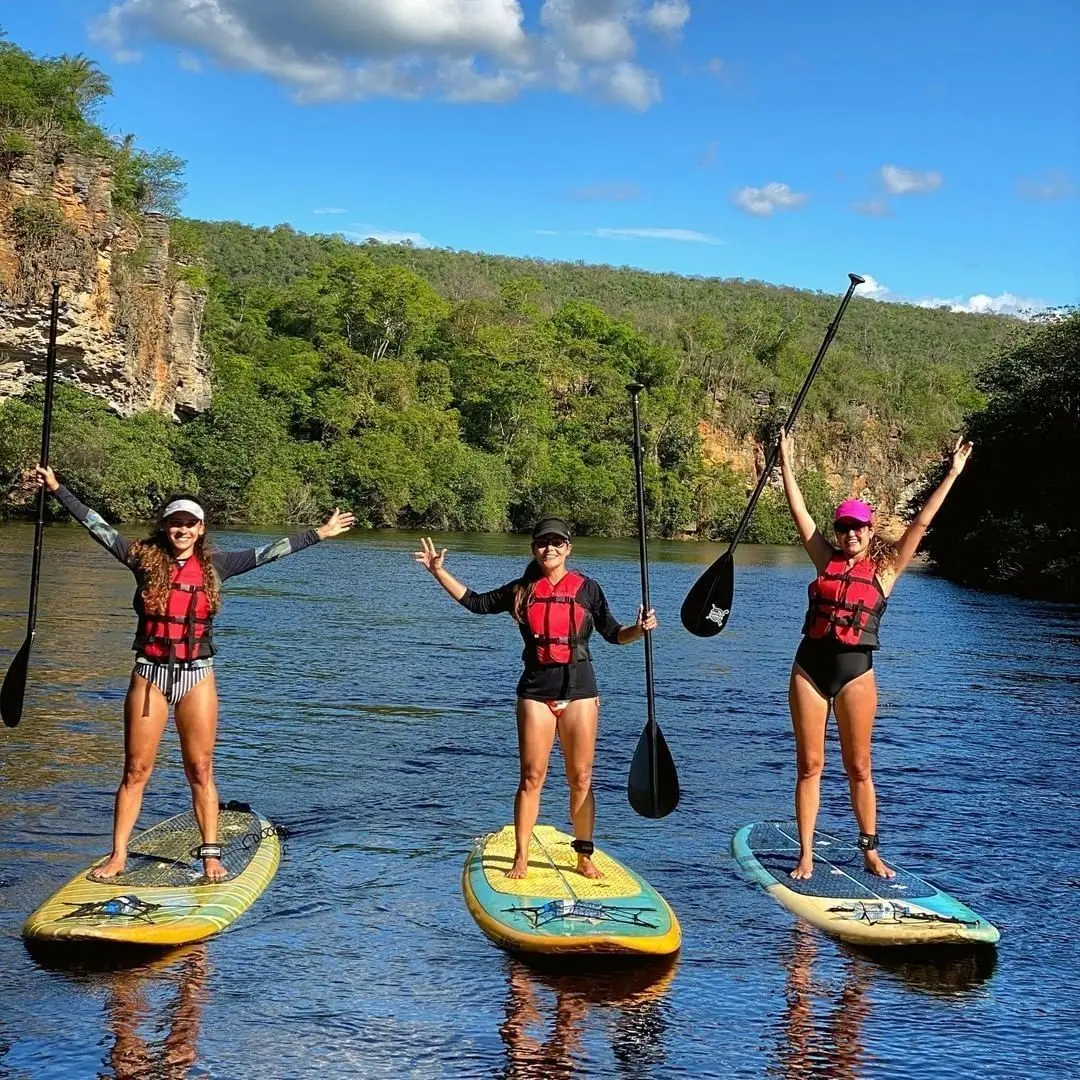 Três mulheres de colete remando em pranchas de stand up num rio calmo entre paredões