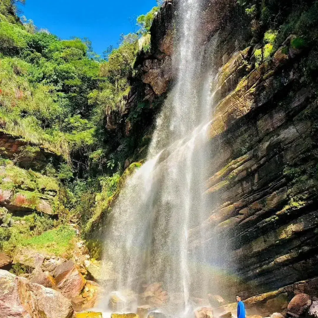 Cachoeira alta descendo um paredão com musgo, com um pequeno arco-íris e uma pessoa de azul na base