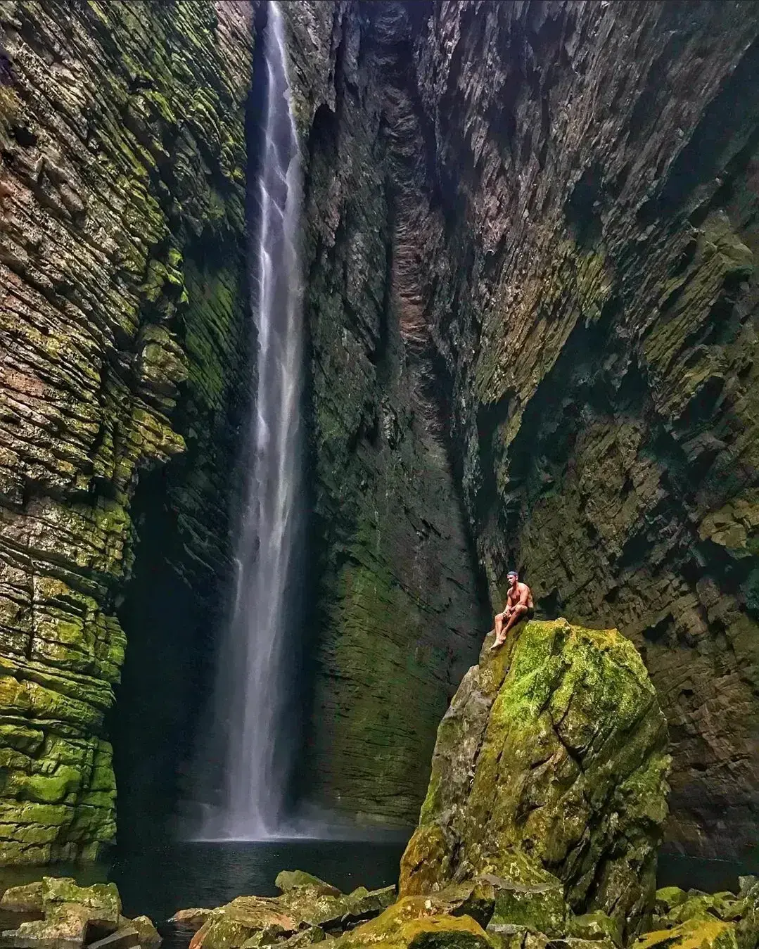 Pessoa sentada numa pedra sob uma cachoeira alta que cai num poço escuro