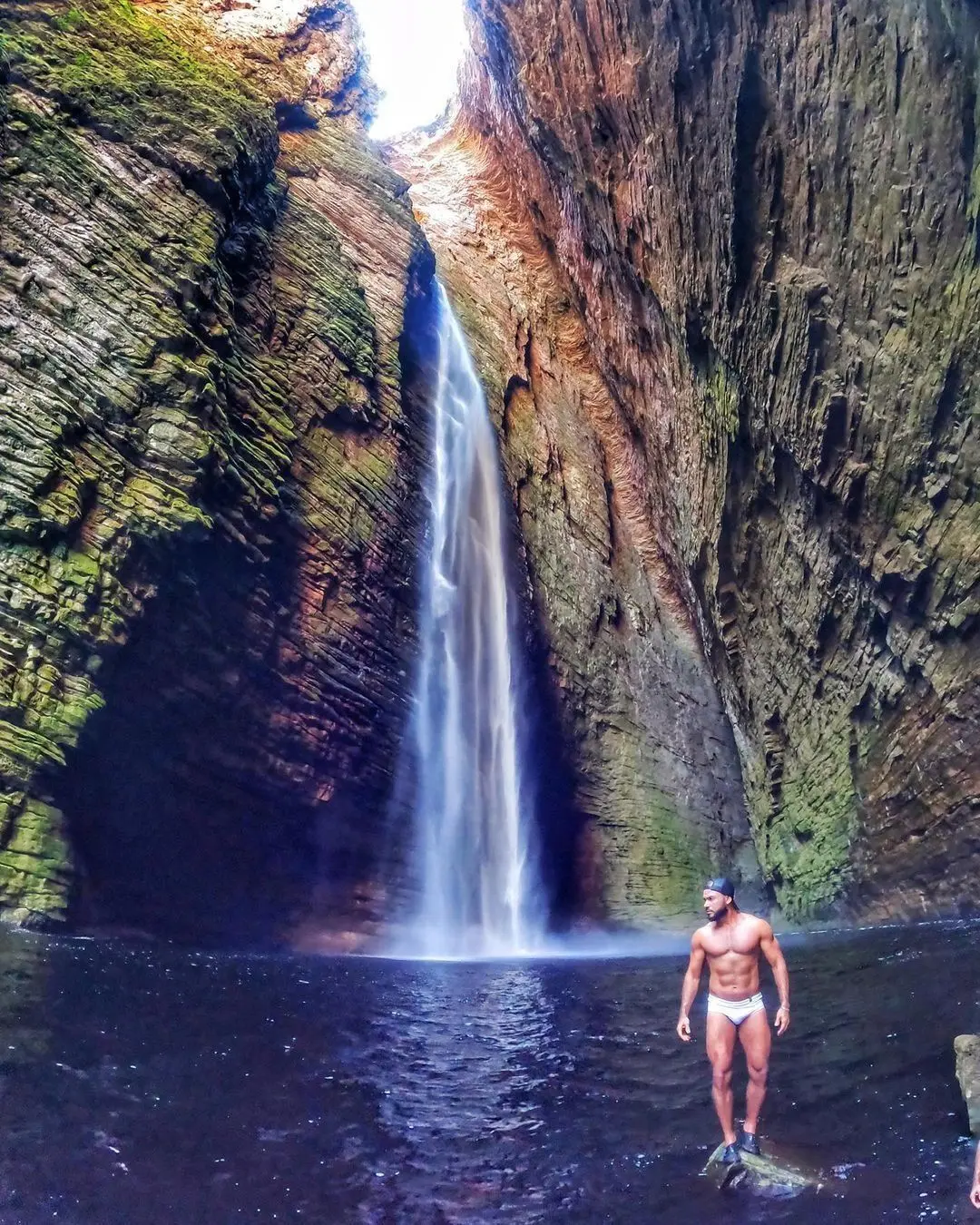 Homem de pé numa pedra no poço diante de uma cachoeira alta que cai por um cânion estreito
