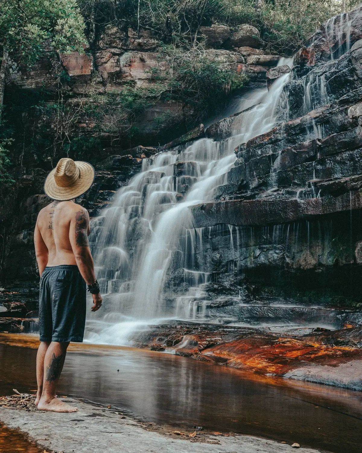 Homem de chapéu de palha diante de uma cachoeira que desce em degraus de rocha escura
