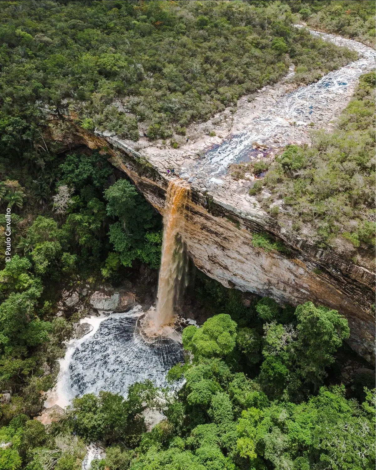 Vista aérea de uma cachoeira cor de chá caindo de uma laje de pedra num poço cercado de mata