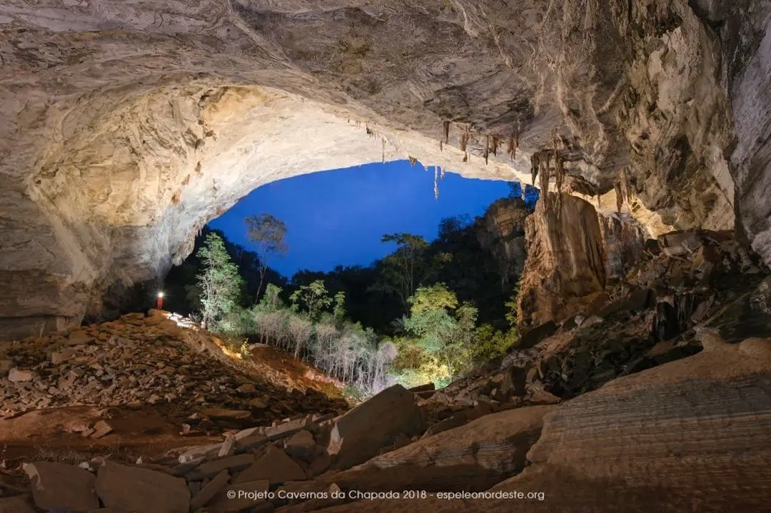 Boca de caverna com estalactites se abrindo para a mata ao anoitecer, com uma pessoa de lanterna ao longe