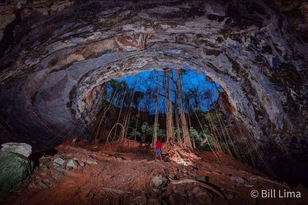 Entrada de caverna com raízes de árvore pendendo do teto e uma pessoa de vermelho