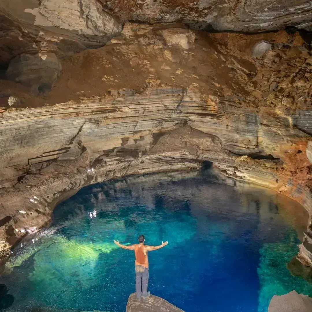 Pessoa de braços abertos diante de um lago azul dentro de uma gruta