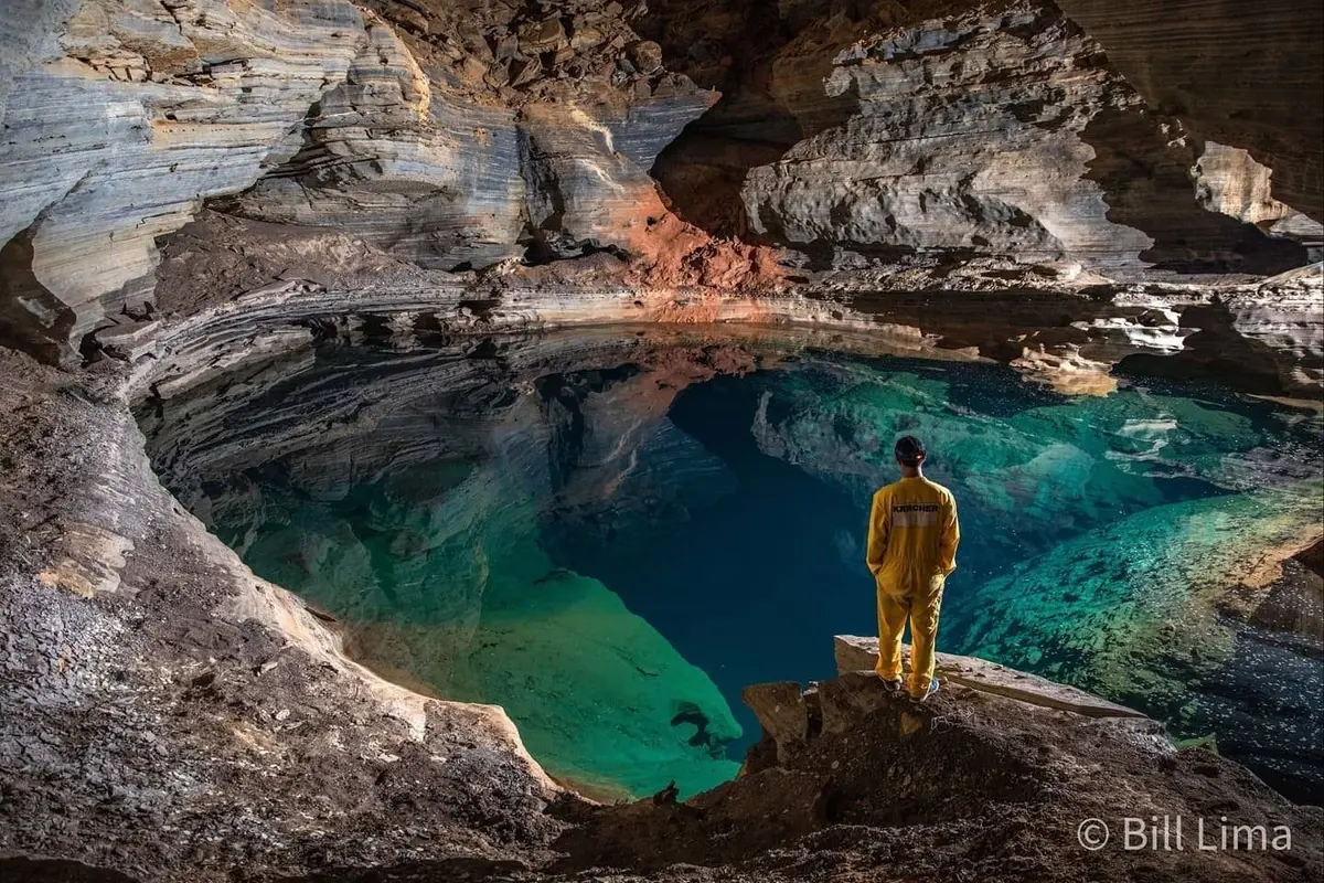 Espeleólogo de macacão amarelo diante de um lago verde-azulado dentro de uma caverna