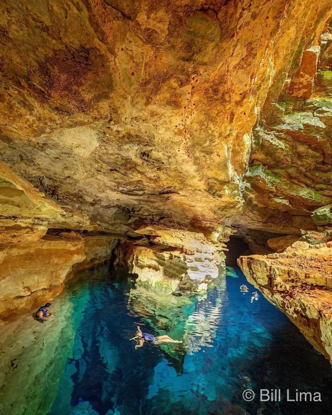Visitantes de colete e máscara flutuando na água azul do Poço Azul, entre paredes alaranjadas