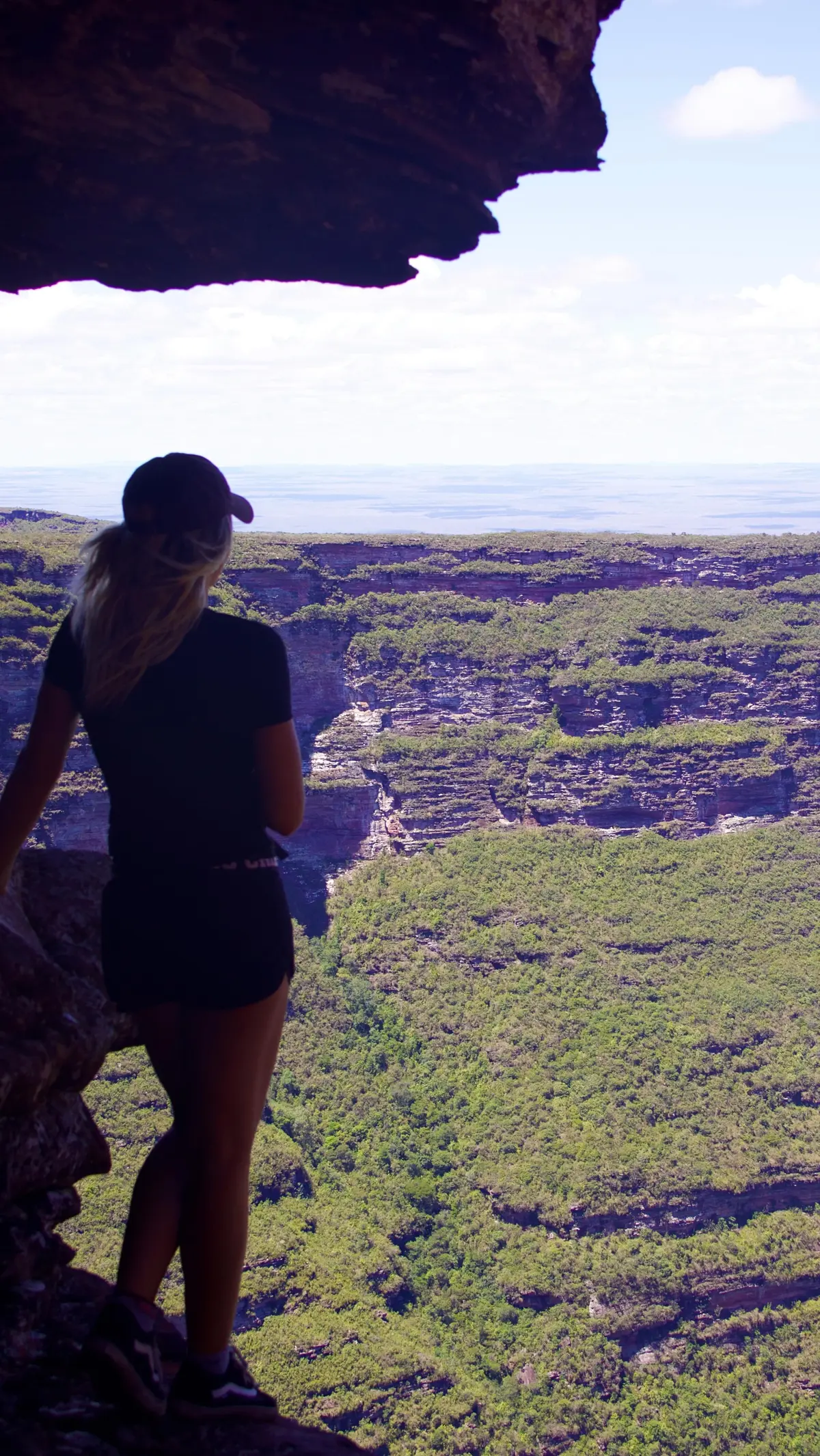 Mulher sob uma laje de pedra olhando um cânion enorme coberto de mata