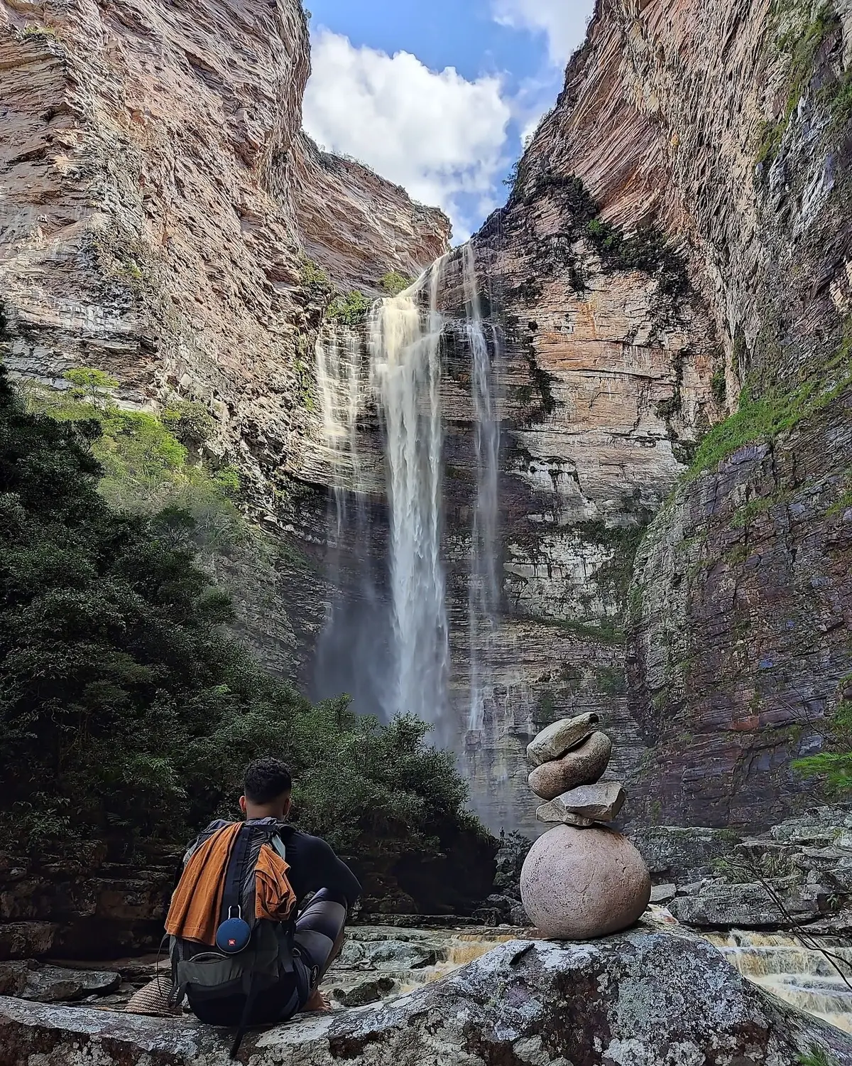 Mochileiro sentado numa pedra diante de uma cachoeira alta num cânion de paredões