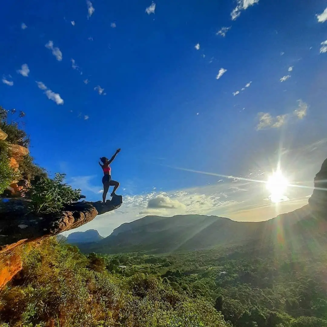 Mulher sentada numa pedra em balanço ao pôr do sol, com vale e serras ao fundo