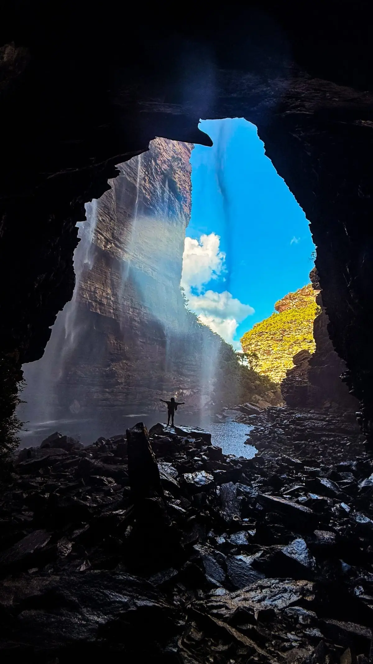 Silhueta de braços abertos dentro de uma gruta, diante de uma cachoeira que cai pelo paredão