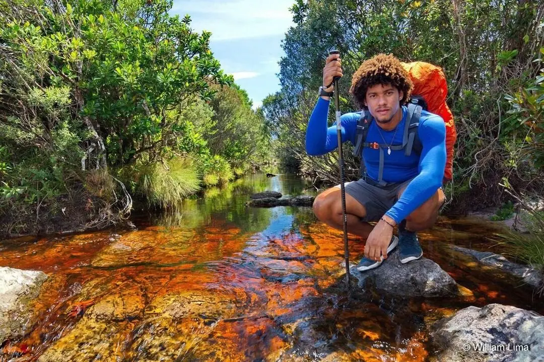 Caminhante com mochila cargueira agachado num riacho cor de chá entre arbustos