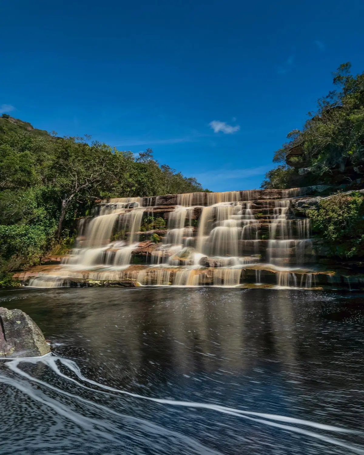 Cachoeira do Riachinho: cascata larga descendo degraus de pedra até um poço escuro, sob céu azul