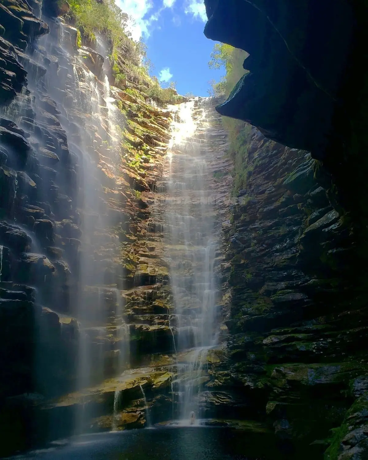 Cachoeira alta caindo entre paredões de rocha em camadas até um poço escuro, vista de dentro do cânion