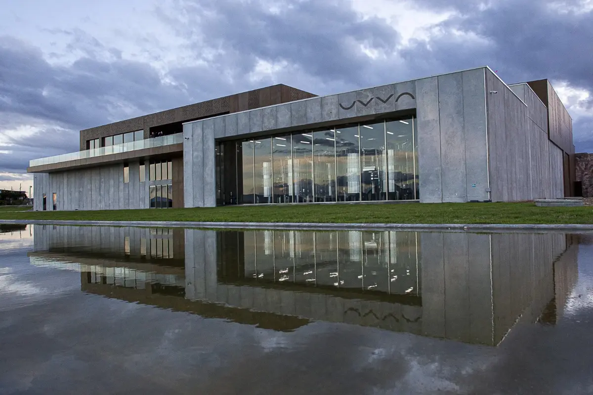 Prédio de concreto e vidro da vinícola UVVA refletido num espelho d’água, sob céu nublado ao entardecer