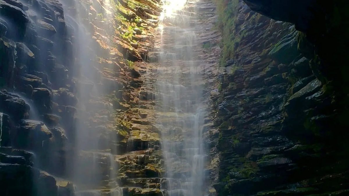 High waterfall falling between layered rock walls into a dark pool, seen from inside the canyon