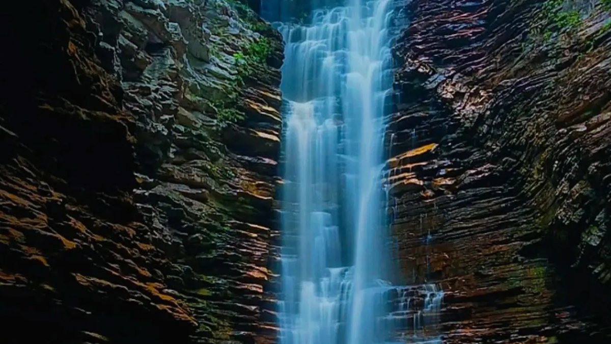Buracão Waterfall: a tall, thin waterfall between layered rock walls, falling into a dark pool under the blue sky